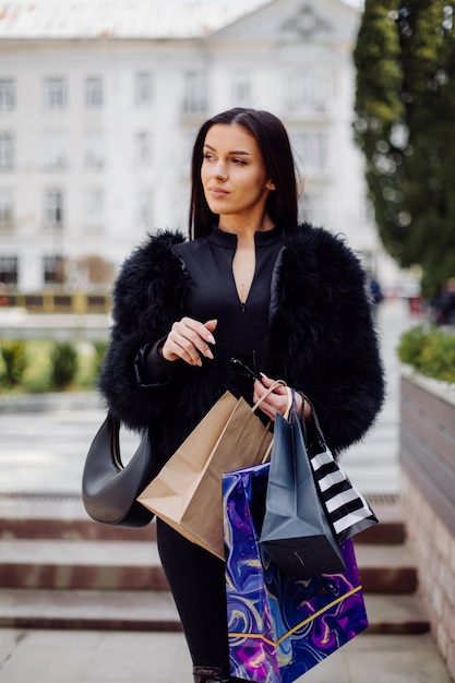 검은 옷을 입은 갈색 머리의 여인이 성공적인 쇼핑을하는 동안 화려한 무늬의 쇼핑백을 들고 있습니다. 밖으로 걸어가는 그녀는 하루의 온기를 즐기고있다