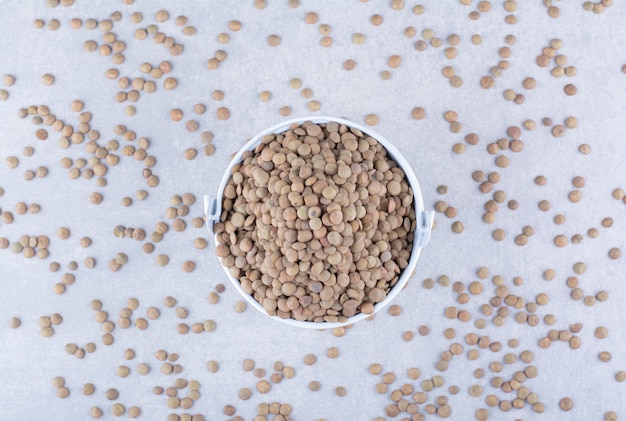 Brown lentil filled into a small bucket sitting in the midst of scattered grains on marble surface