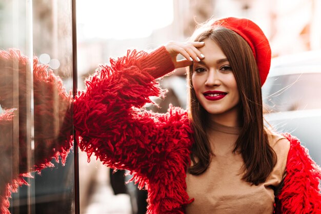 Browneyed attractive woman in great mood posing on street Closeup shot of fashionable girl with red lipstick dressed in bright coat hat and beige Tshirt