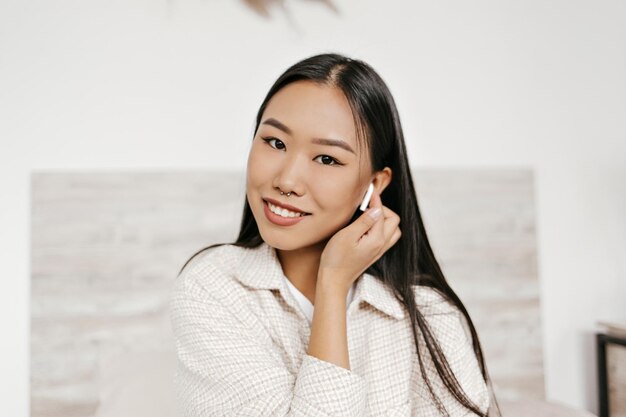 Browneyed brunette Asian woman in good mood listens to music in wireless headphones and smiles sincerely Portrait of attractive lady in beige jacket looks into camera in white room