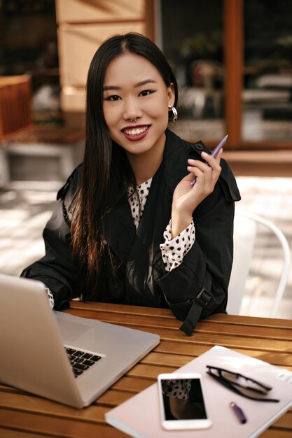 Browneyed brunette woman smiles sincerely looks into camera works with laptop and sits at wooden desk outside