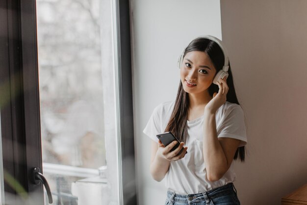 Browneyed girlはカメラを見て、ヘッドフォンで歌を楽しんでいる白いトップの電話アジアの女性を保持します