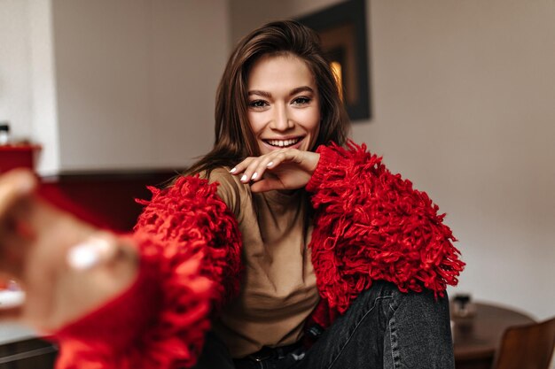 Browneyed smiling girl dressed in jeans top and red cardigan makes selfie sitting in kitchen