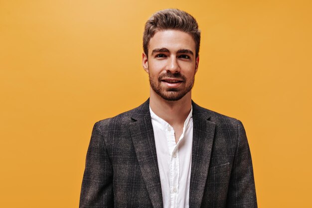 Browneyed stylish brunet bearded man in white shirt and checkered jacket smiles and looks into camera on orange background