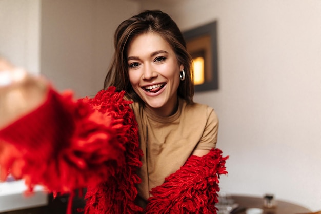 Browneyed woman in beige top makes selfie Brunette in red knitted jacket shows tongue