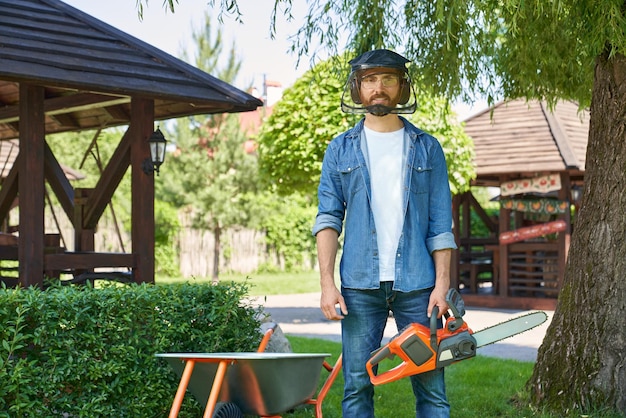 Free photo brunet handyman in protective shield standing with cordless chain saw while working in backyard