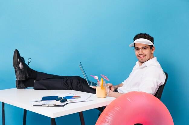 帽子と事務服を着たブルネットの男は、テーブルの上に足を置いて座っています。ガイはラップトップを持って、インフレータブルサークルのある孤立した空間でカクテルを楽しみながら仕事をしています。