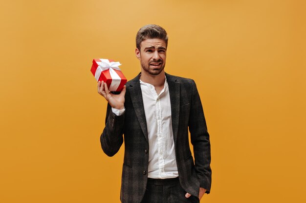 Brunet young bearded man in stylish checkered jacket and white shirt grimaces and holds red gift box on orange background