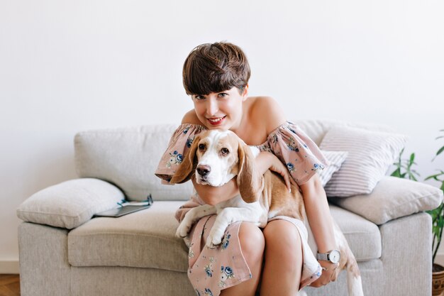 Brunette amused woman wears wristwatch and dress with floral ornament holding big puppy on her knees and smiling