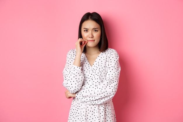 Free photo brunette asian woman in dress looking displeased, frowning and zipping mouth, seal lips with promise, standing over pink background.