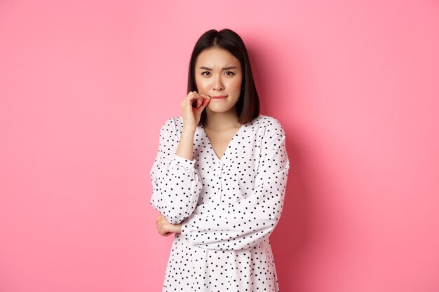 Brunette asian woman in dress looking displeased, frowning and zipping mouth, seal lips with promise, standing over pink background.