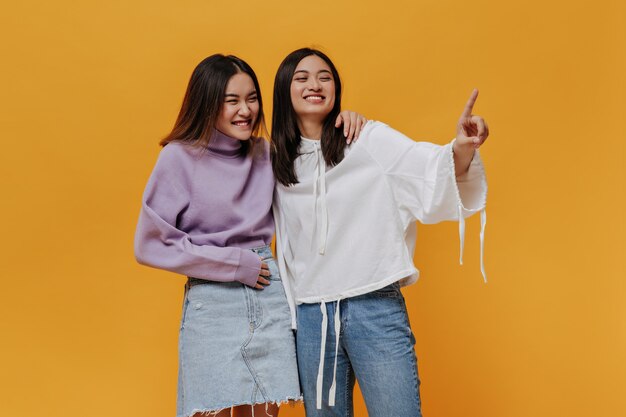 Brunette Asian woman in jeans and white hoodie smiles, hugs her girlfriend and points at place for text on isolated orange wall