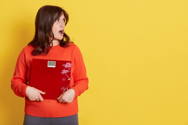 Brunette athletic woman holding scale and expressing surprise on face, looking aside with opened mouth, model posing over yellow, copy space for advertisement.