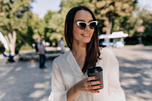 Brunette attractive woman in elegant white blouse is smiling and looking aside on park background