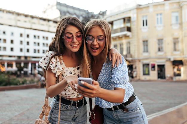 세련된 의상을 입은 갈색 머리와 금발의 여성들은 놀란 표정을 짓고 휴대폰으로 메시지를 읽습니다.