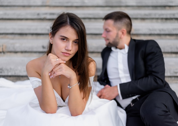 Brunette bride with big brests is looking straight and a groom is sitting on the background