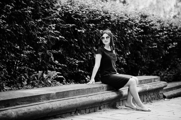 Brunette business girl at black dress on sunglasses sitting at border against bushes posing at street of city