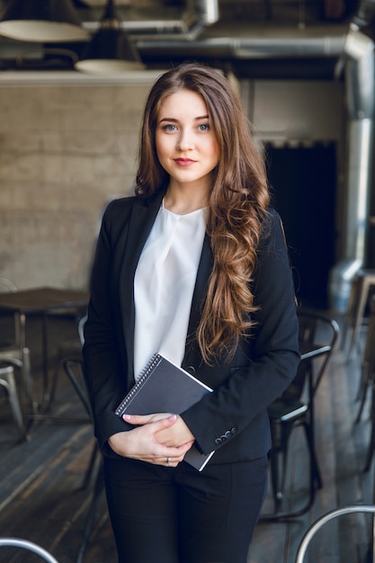 Brunette business woman with wavy long hair and blue eyes stands holding a notebook in hands
