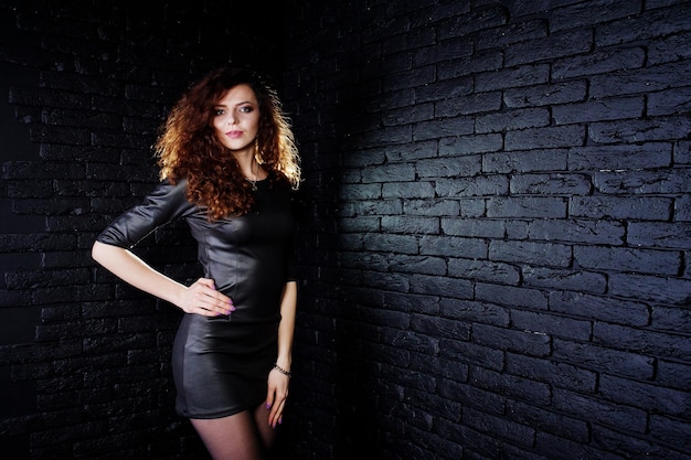 Brunette curly haired long legs girl in black leather dress posed at studio against dark brick wall