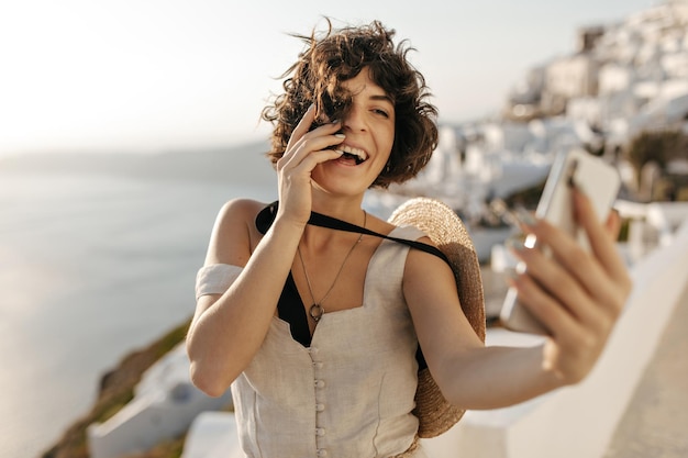 Bruna donna riccia in abito beige e cappello di paglia sorride sinceramente e si fa selfie fuori nella vecchia città greca felice signora in abito elegante e barcaiolo tiene il telefono sullo sfondo del mare