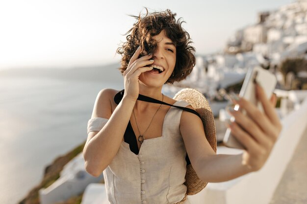 Брюнетка кудрявая женщина в бежевом платье и соломенной шляпе искренне улыбается и делает селфи на улице в старом греческом городе Счастливая дама в стильном наряде и канотье держит телефон на фоне моря