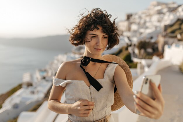 Brunette curly woman in beige dress and straw hat takes selfie on sea and city wall