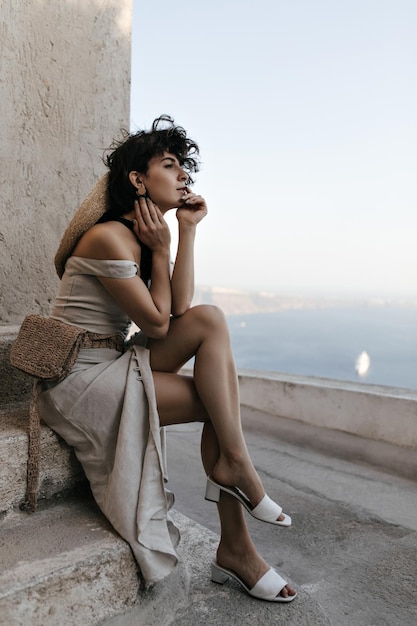 Free photo brunette curly woman in beige dress with straw bag enjoys sea view young lady in stylish outfit sits near old house