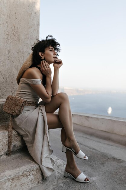 Brunette curly woman in beige dress with straw bag enjoys sea view Young lady in stylish outfit sits near old house