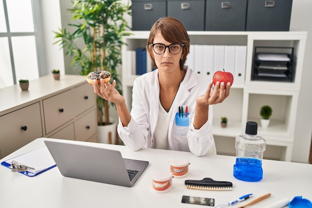 Foto gratuita bruna dentista donna con mela e ciambella al cioccolato scettico e nervoso, accigliato sconvolto a causa del problema. persona negativa.