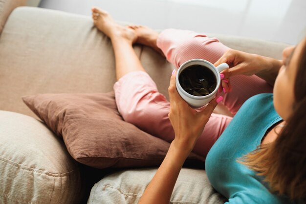 Брюнетка в уютной домашней одежде держит чашку чая, смотрит в окно и отдыхает на современном диване