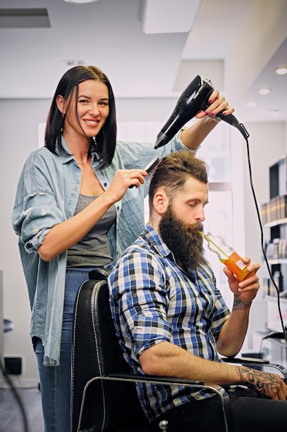 Brunette female hairdresser doing a hairstyle with hair dryer of the bearded men client in a saloon.
