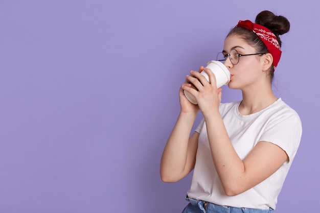 Foto gratuita la donna castana tiene e beve caffè per andare, gode di un drink piacevole, indossa una maglietta casual bianca e una fascia per capelli, si concentra da parte, gode di bevande calde