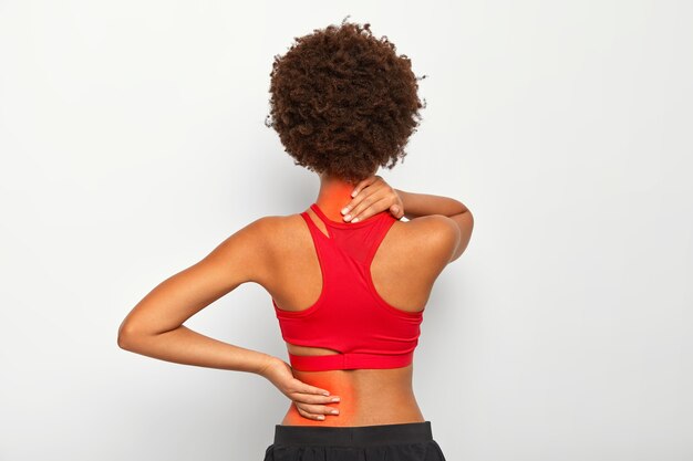 Brunette female model has joint pain in back and neck, has health problem, dressed in active wear isolated over white studio wall