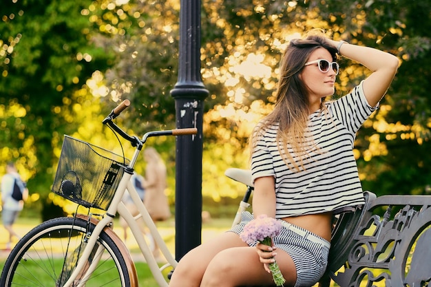 Brunette female sits on a bench and holds flower bouquet with bicycle in a park on background.