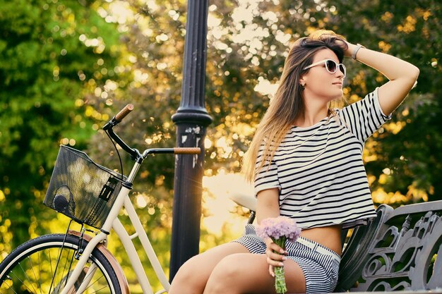 Brunette female sits on a bench and holds flower bouquet with bicycle in a park on background.