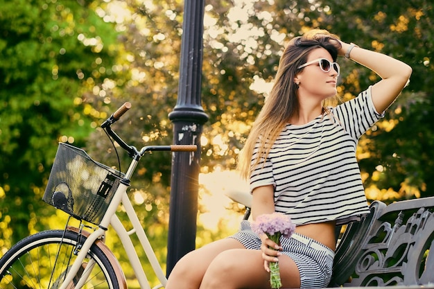 Brunette female sits on a bench and holds flower bouquet with bicycle in a park on background.