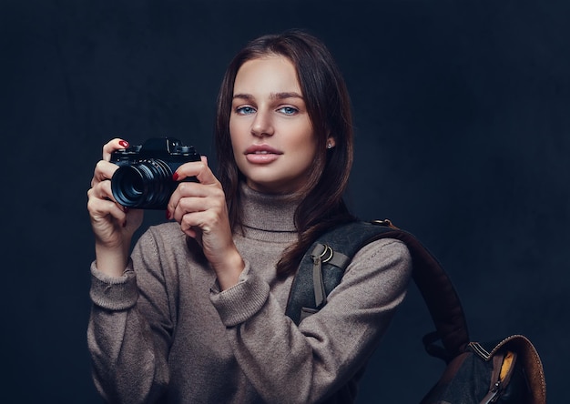 A brunette female traveler with backpack holds compact photo camera.