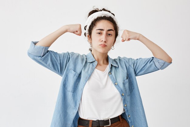 Brunette female wearing do-rag and denim shirt, shows muscles and strength, has confident expression, ready to fight or protect herself. Cute tender woman has power, being sure in victory.