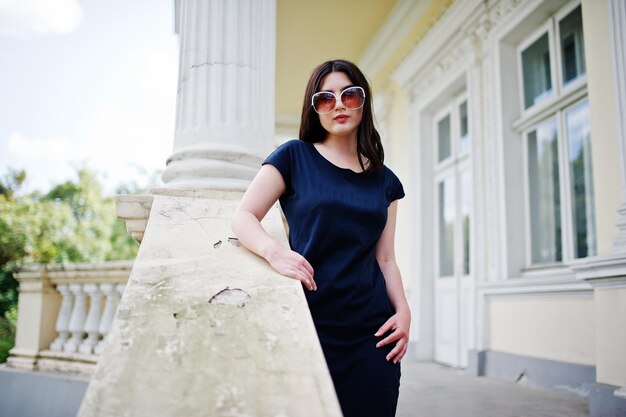 Brunette girl at black dress sunglasses posed against old vintage house at street of city