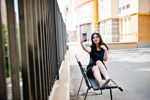 Brunette girl at black dress sunglasses sitting on bench listening music from headphones phone and posing at street of city