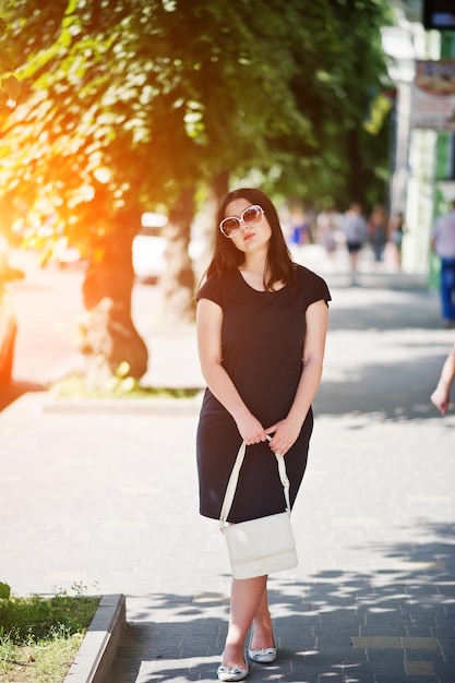 Brunette girl at black dress on sunglasses with handbag at hand posing at street of city