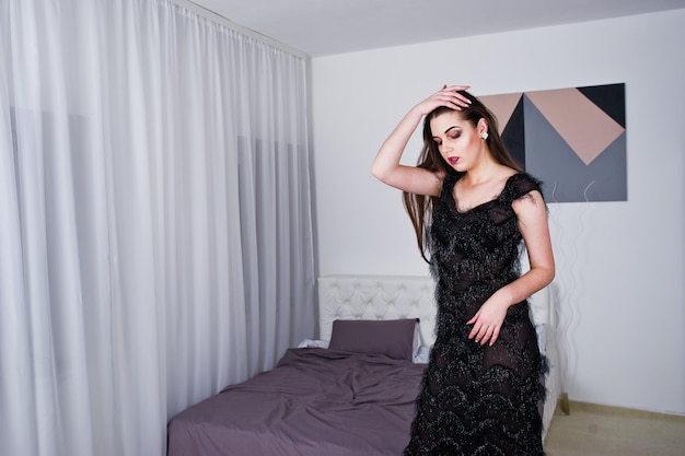 Free photo brunette girl in black evening dress against white room with bed