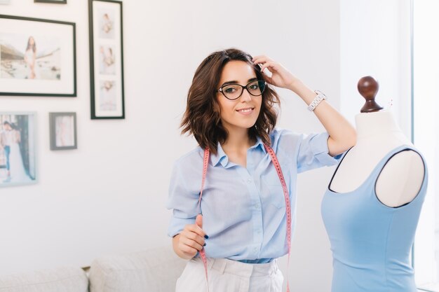 Brunette girl in blue shirt standing near a mannequin in a workshop studio. She creates a blue dress . She is smiling at the camera.