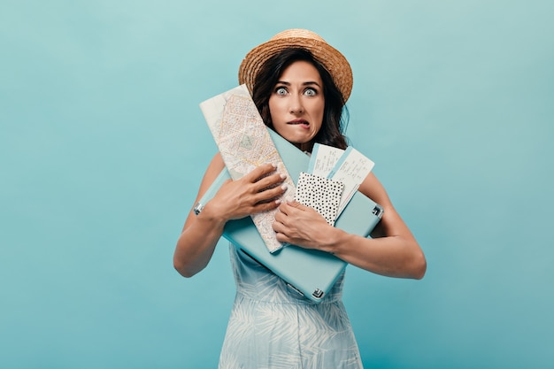 Brunette girl feels awkward and poses with suitcase, tickets on blue background. Woman in straw hat with map in her hands and in blue dress.