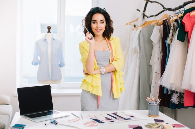 A brunette girl in a gray dress and yellow jacket is standing near the table in a workshop studio. She has a lot of creative stuff  on the table . She is holding a pencil in a hand.