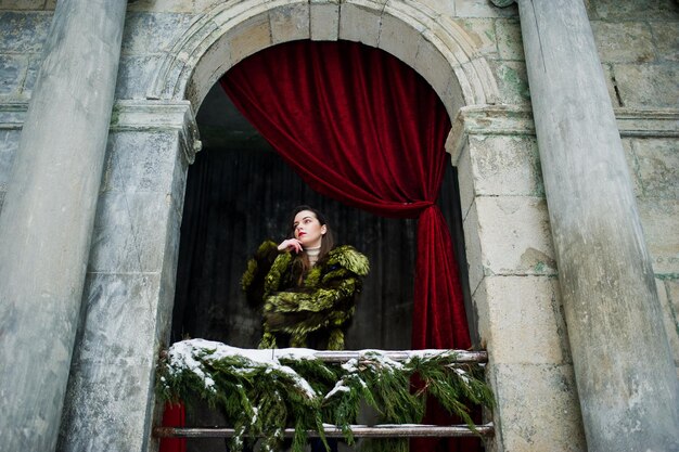 Brunette girl in green fur coat against old arch with columns and red curtains