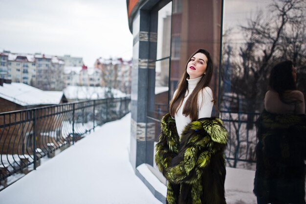 Brunette girl in green fur coat at street of city against house with large windows at winter