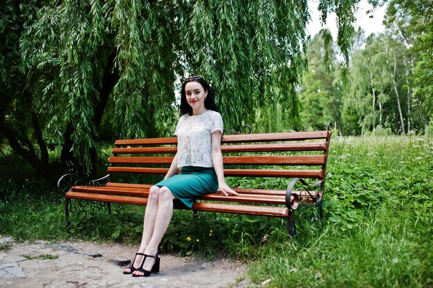 녹색 치마와 흰색 블라우스에 갈색 머리 소녀는 벤치에 앉아 공원에서 포즈