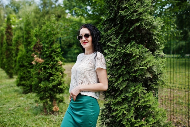Брюнетка в зеленой юбке и белой блузке с солнцезащитными очками позирует в парке