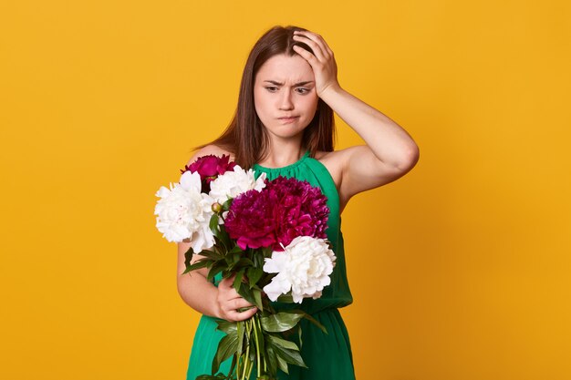 brunette girl holds big bouquet of burgundy peonies in hand, thoughtful woman with flowers keeps hand on head, poses on yellow. Copy space for promotion